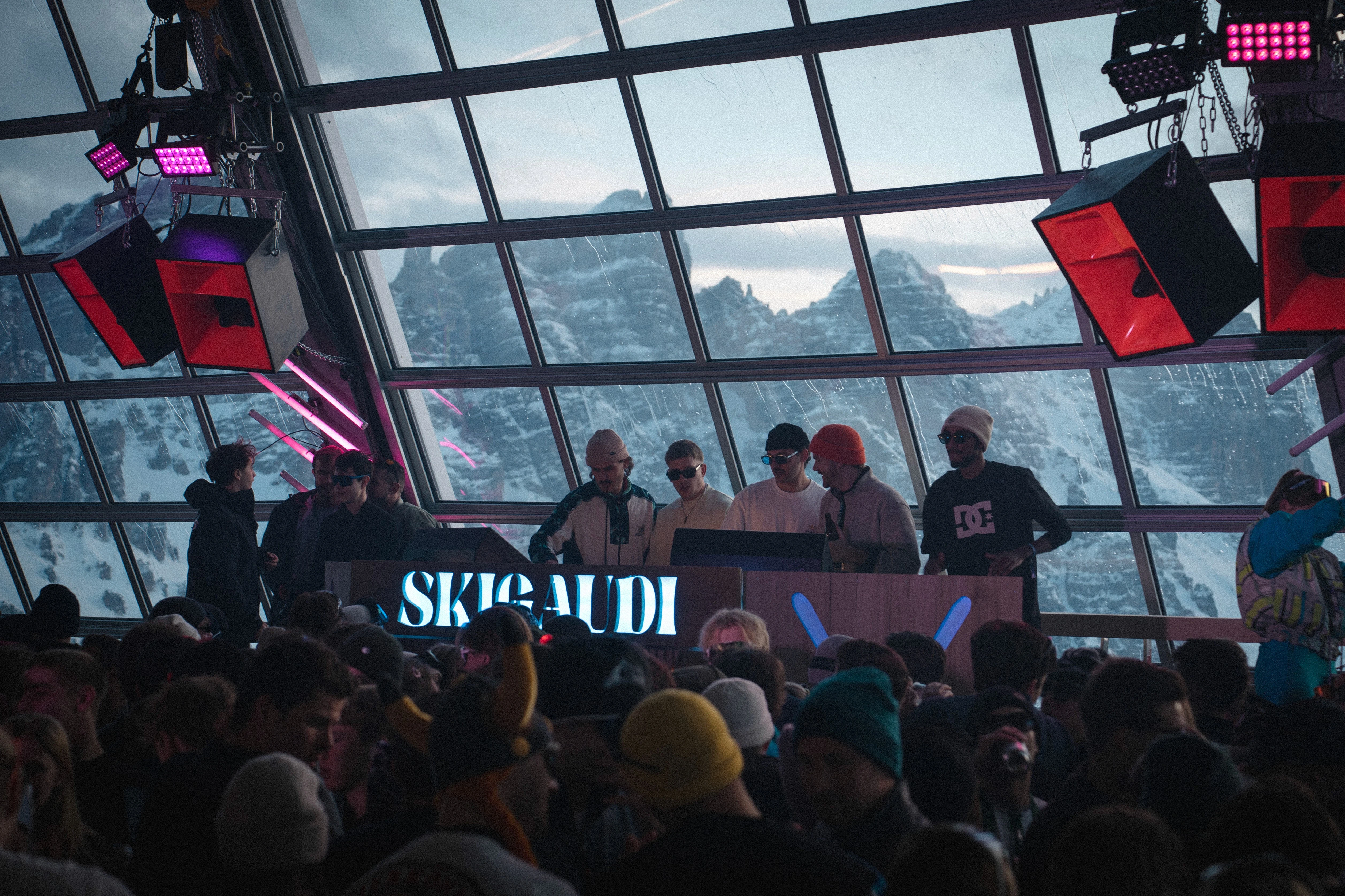 Indoor ski party with DJs and crowd, snowy mountain backdrop.