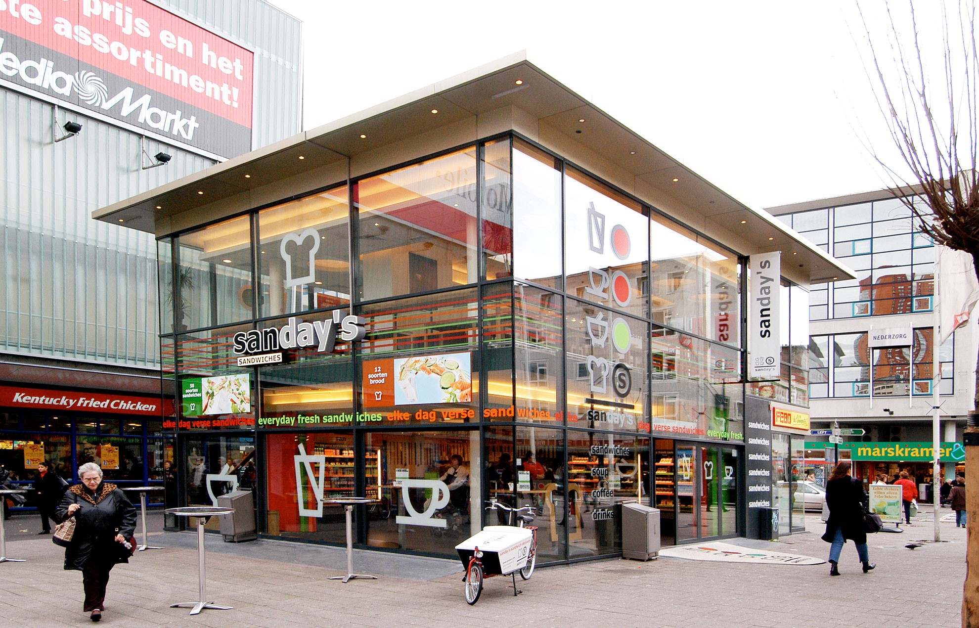 Buitenkant van de sandwichzaak Sanday's in Rotterdam, met een modern glazen ontwerp, menu-symbolen op de ramen, en voorbijgangers op straat.