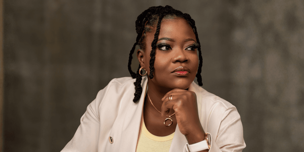 A woman with braided hair gazes thoughtfully to the side, resting her chin on her hand. She wears a light blazer, a necklace, and a confident expression.