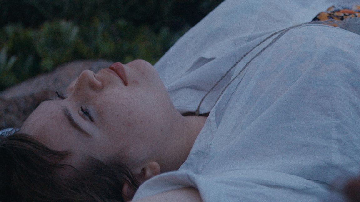 A person lies down outdoors, gazing upward with a serene expression against a natural background.