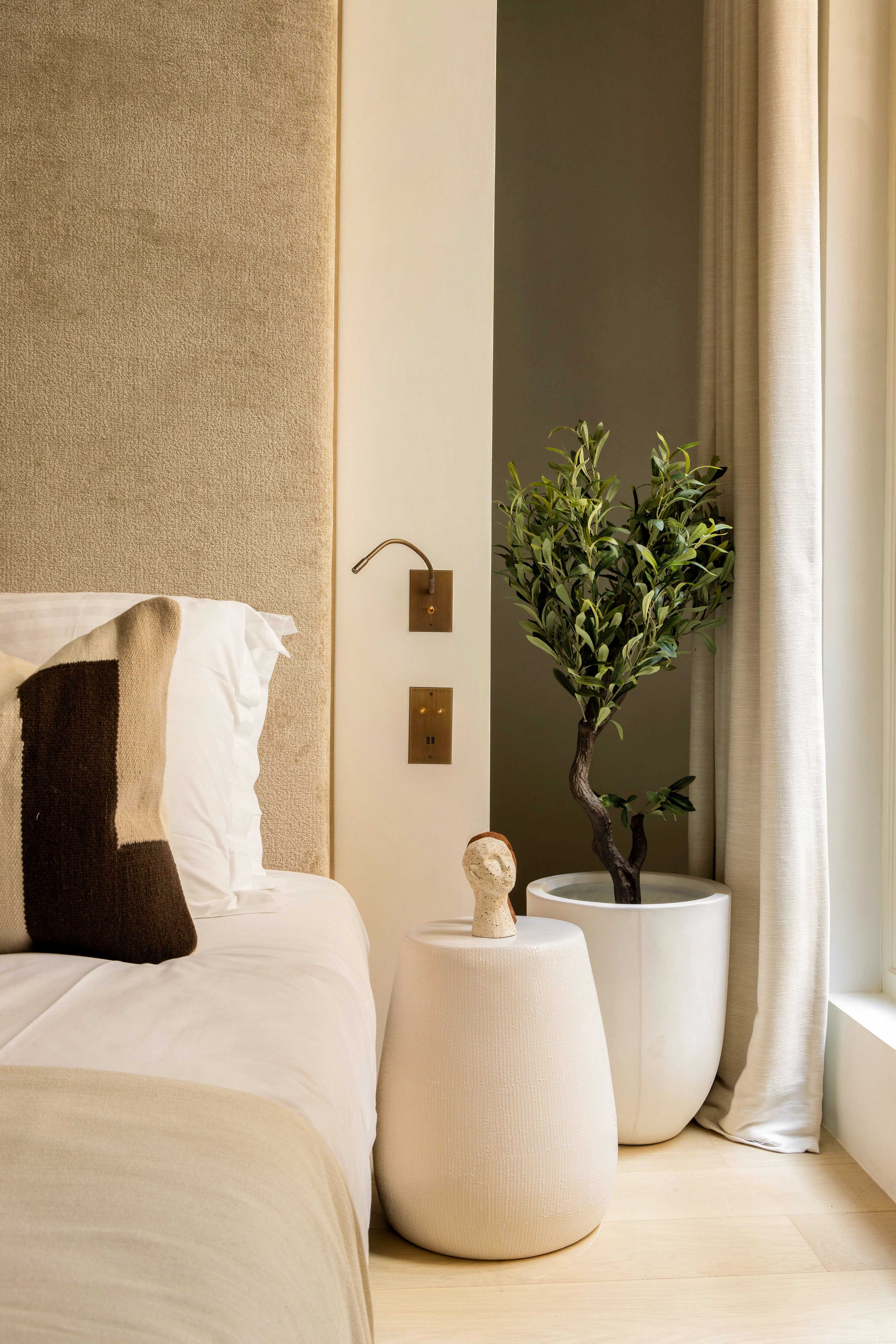 Detail chevet avec table d'appoint ceramique blanche, liseuse doree, petit olivier et tete de lit velours sable