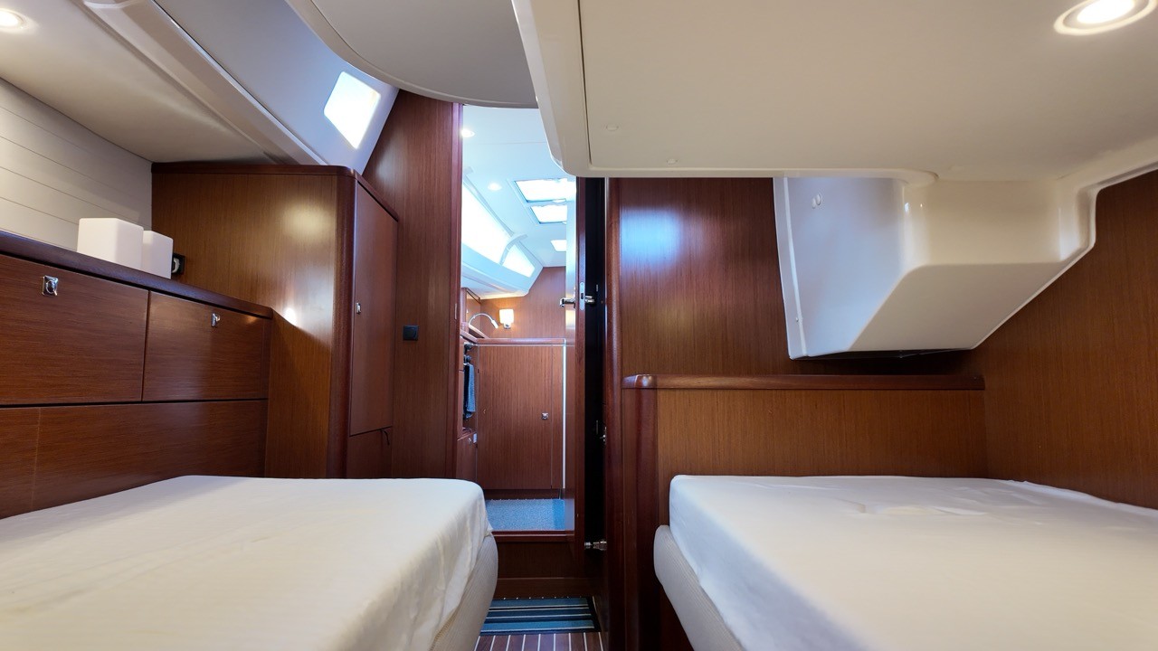 Beneteau Clipper 361 main saloon interior showing the stainless steel galley sink with chrome tap, white worktop, mahogany cabinetry throughout, nav station with red panel, analogue gauges and chartplotter, teak-and-holly cabin sole, blue fabric U-shaped dinette seating with folding table, and a flat-screen TV mounted in the forward bulkhead