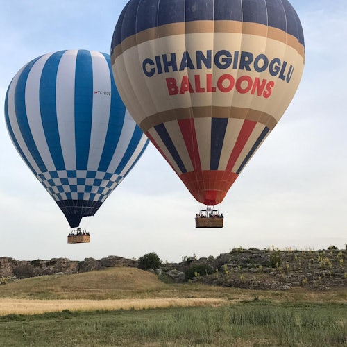 Dalawang hot air balloons, ang isa ay may asul na guhit at ang isa pa ay may marka na "CIHANGIROGLU BALLOONS," ay lumilipad sa ibabaw ng isang madamo at mabatong tanawin.
