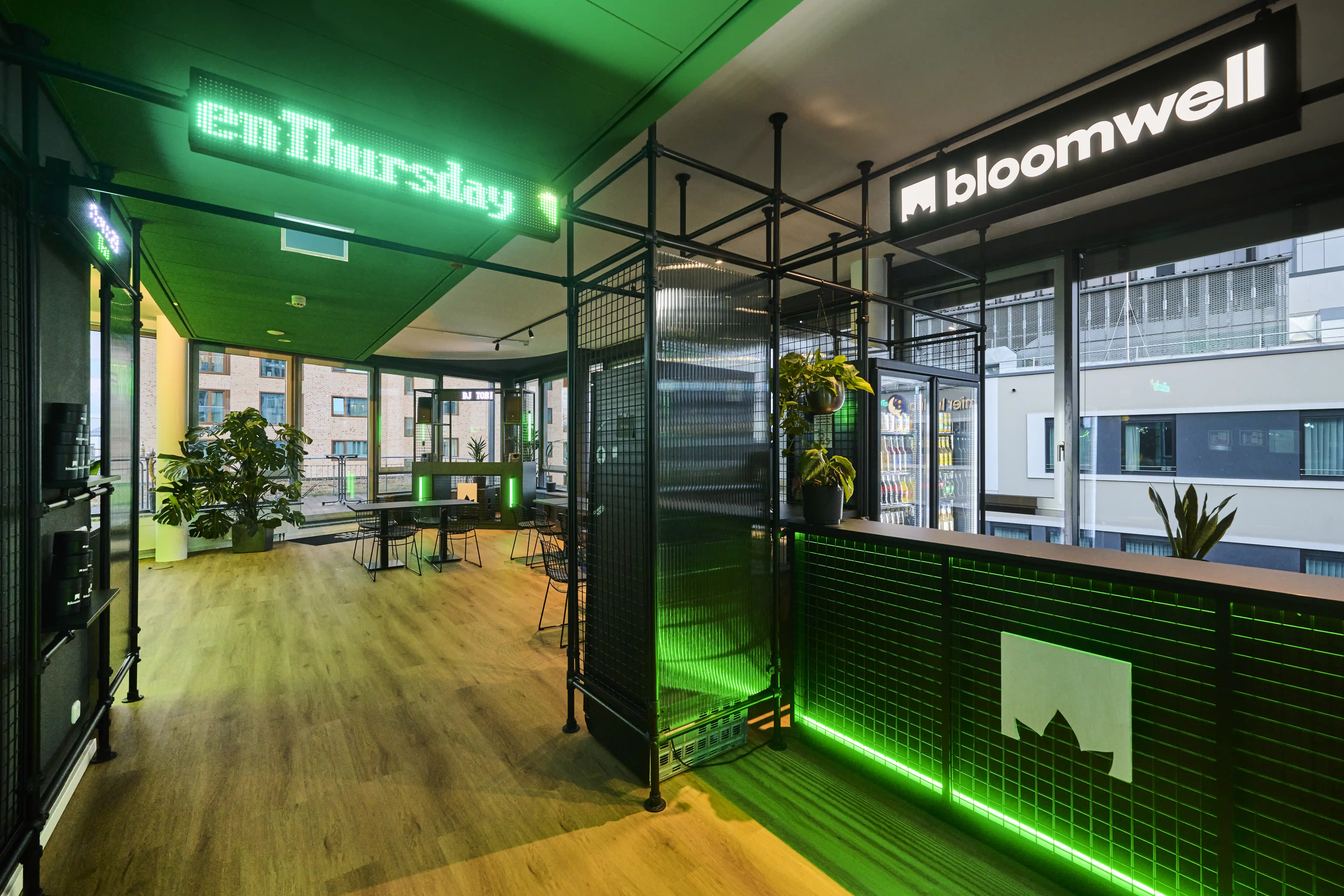 Entrance at BLOOMWELL with a black mesh reception counter lit by green LED strips, a leaf logo, a green enThursday neon sign on the green ceiling and the white bloomwell logo on black.