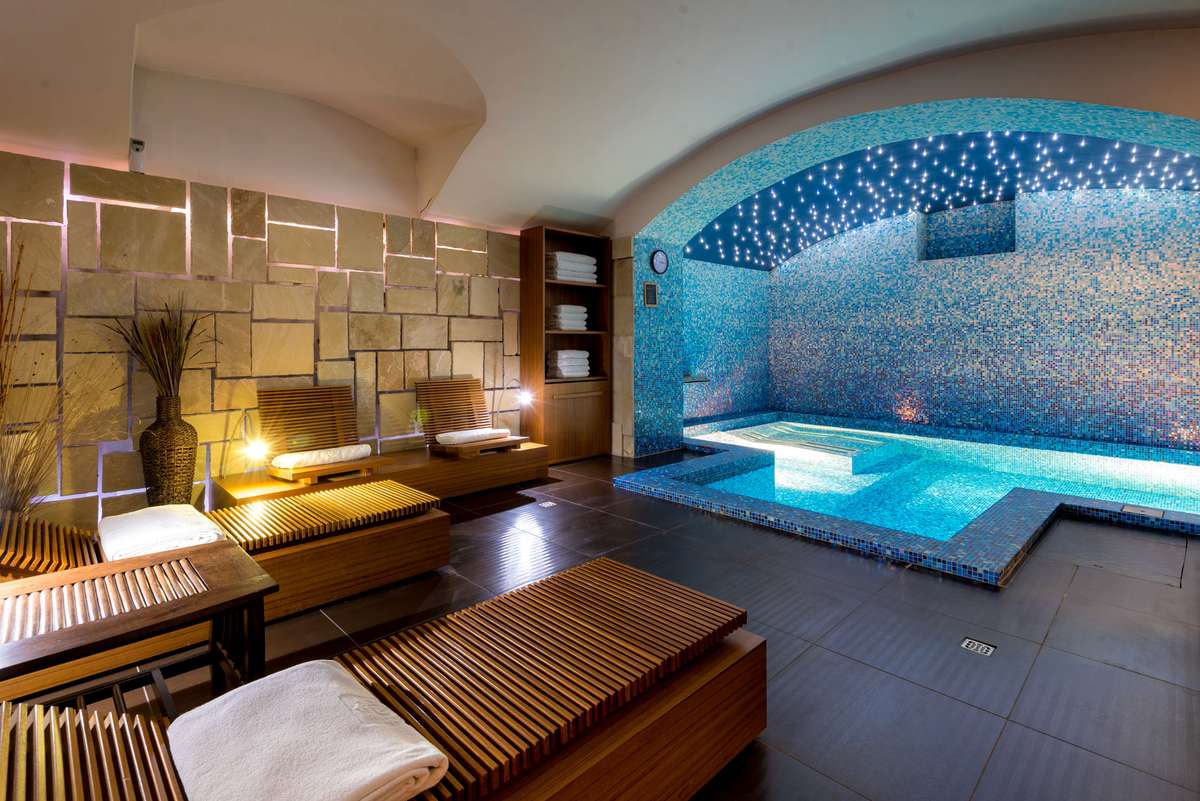 A spa interior features wooden lounge chairs with white cushions and a blue tiled pool under an arched ceiling.