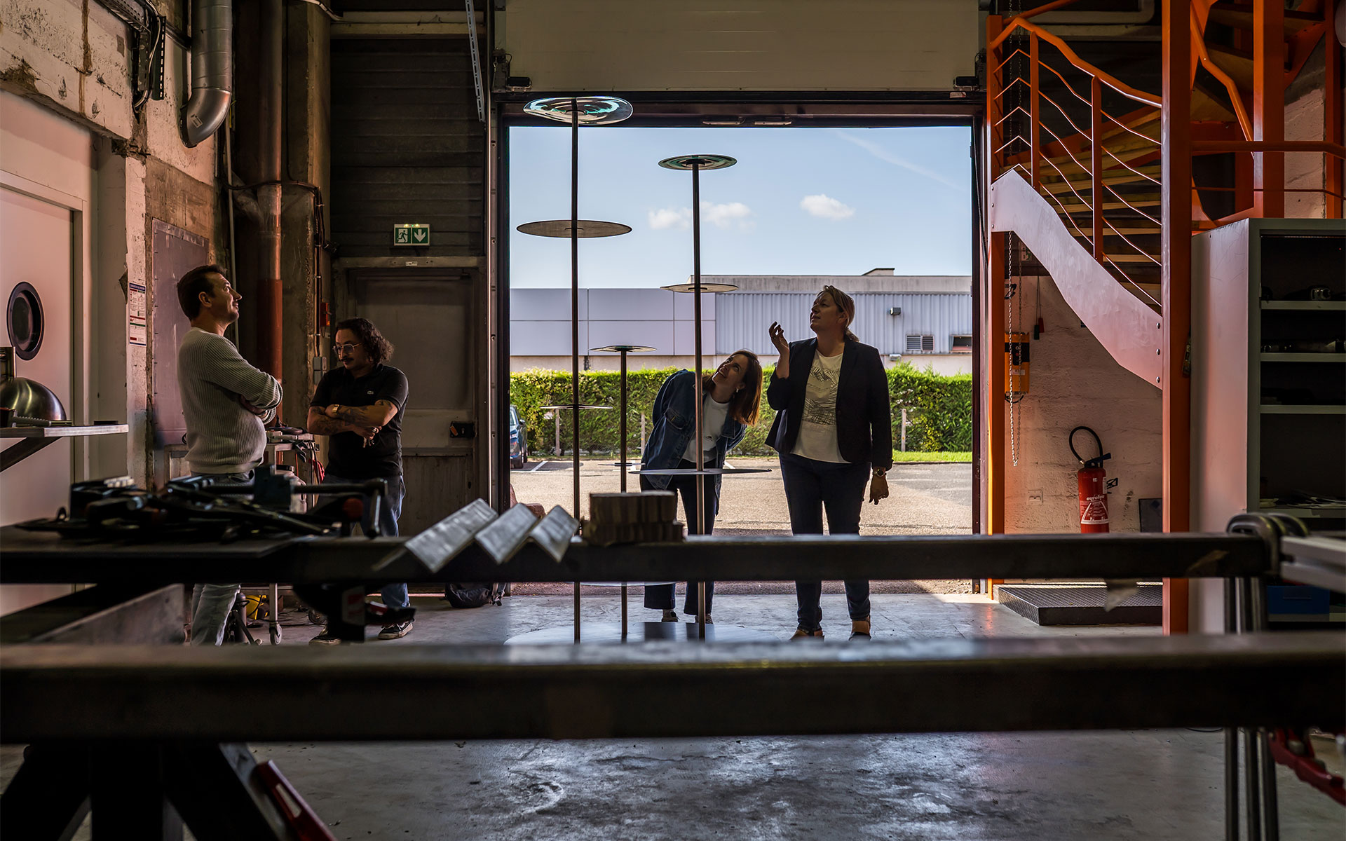 Dans un atelier industriel, quatre personnes sont réunies devant une grande porte ouverte donnant sur l'extérieur. Deux personnes sont situées à gauche dans l'ombre de l'atelier, tandis qu'une femme en veste en jean et une femme en blazer sombre se tiennent plus près de la lumière, examinant des structures verticales et fines qui s'élèvent vers le haut. L'espace de travail est en arrière-plan, avec un escalier orange visible sur le côté droit, créant une ambiance de collaboration professionnelle dans un environnement de fabrication.