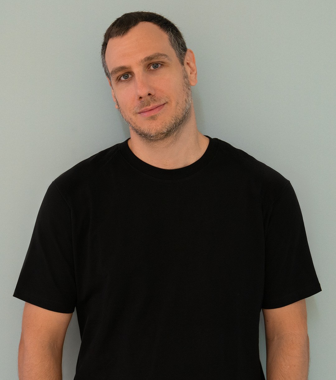 Nik Susic, a creative director, standing against a pale teal wall in a black T-shirt.