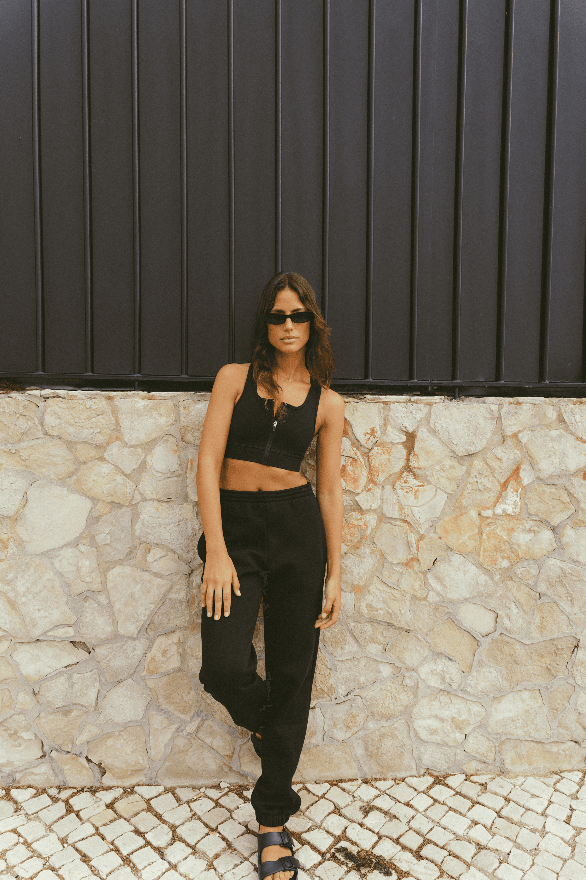 Woman-wearing-a-black-cropped-tank-top-and-black-jogger-pants-standing-against-a-stone-wall-with-one-leg-slightly-bent-and-wearing-black-sandals