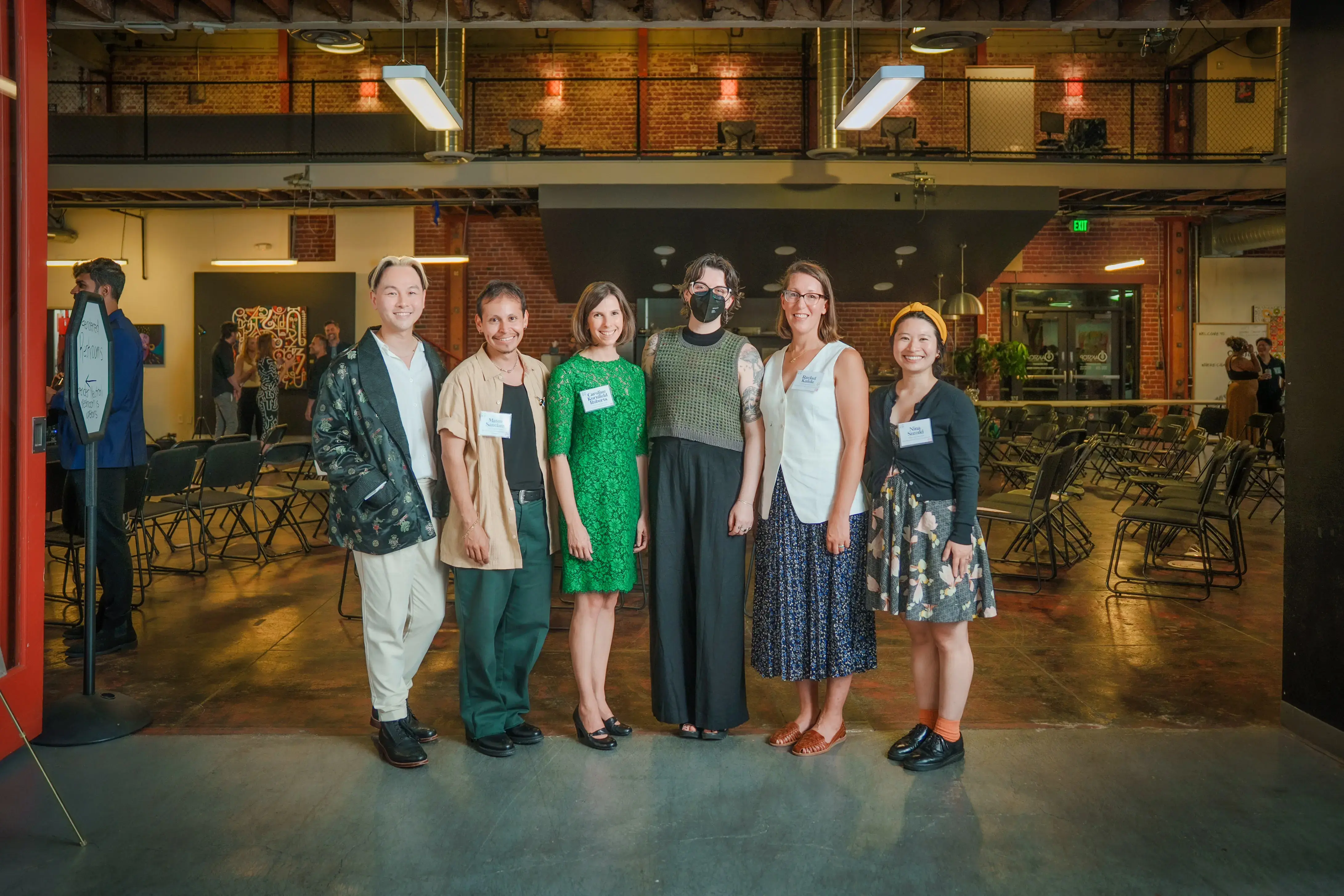 Um grupo de seis pessoas está reunido em um local amplo, de estilo industrial, com paredes de tijolos e vigas expostas, vestidas com uma mistura de trajes casuais e formais, algumas usando crachás, sugerindo um evento profissional ou de networking.