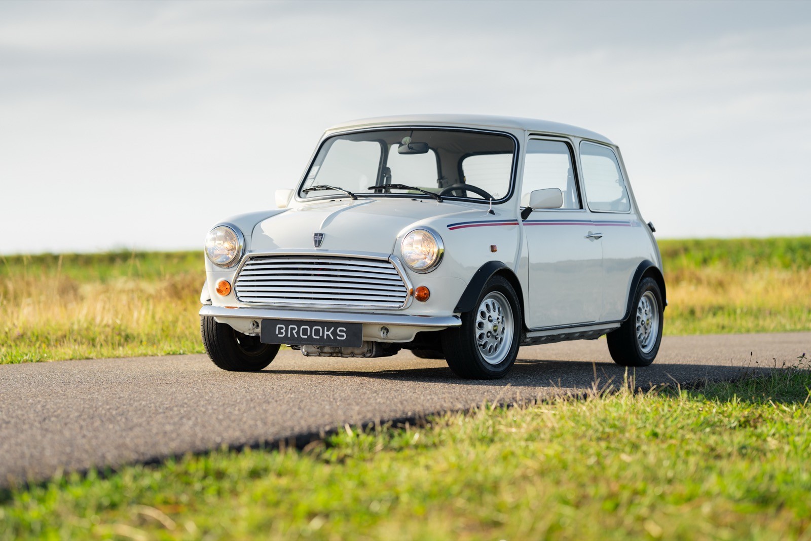 A vintage white car drives along a scenic road surrounded by open grassy fields under a cloudy sky.