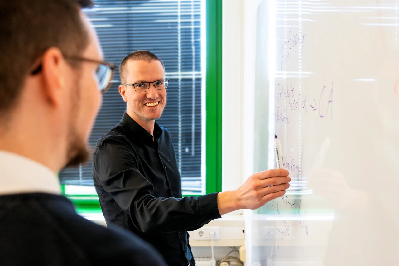 Zwei Männer in einem hellen Büro; einer steht lächelnd vor einem Whiteboard und hält einen Marker, während ein Kollege im Vordergrund ihm zuhört.