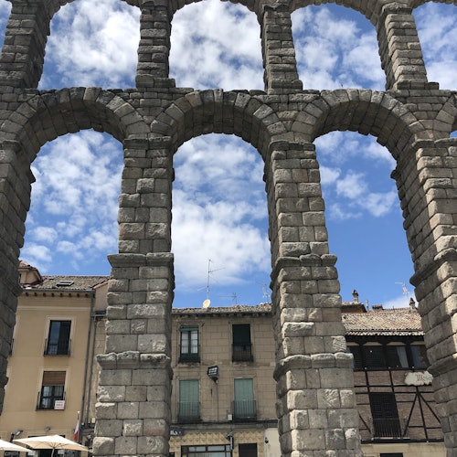 Ancient stone archway with multiple arches in front of old buildings under a blue sky with scattered clouds.