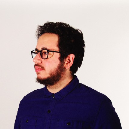 A man with dark curly hair and a beard wears black-framed glasses and a dark blue button-down shirt. He faces left against a plain white background.