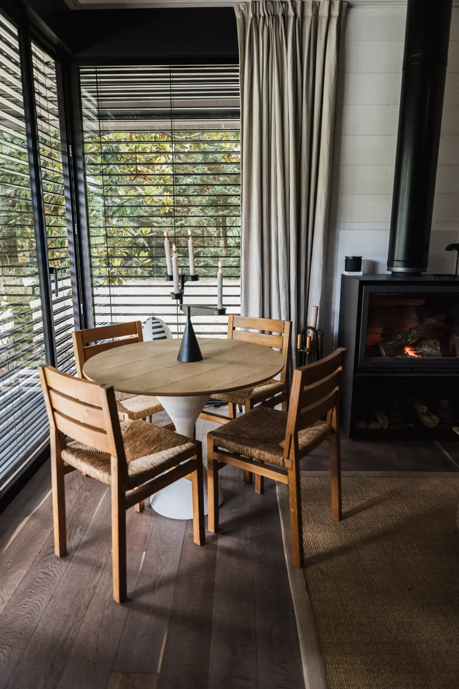 Coin repas du Teshima Bourgogne avec table ronde en bois, chaises paillées, stores à lames et poêle à bois