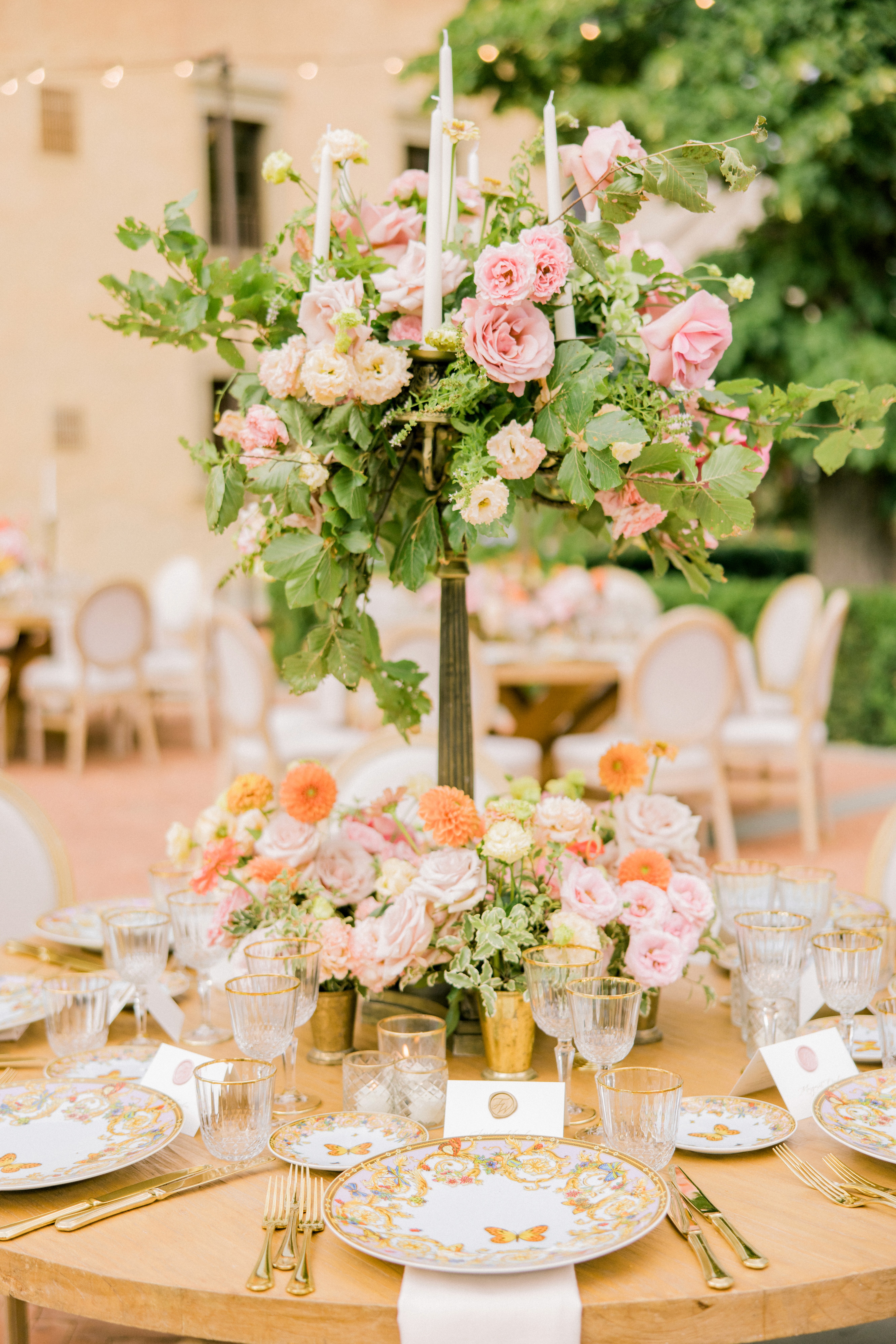 Close-up di tavolo rotondo allestito da luxury catering per matrimonio, con piatti Versace floreali e decorazioni a farfalla, candelabri e fiori coordinati per una mise en place elegante.