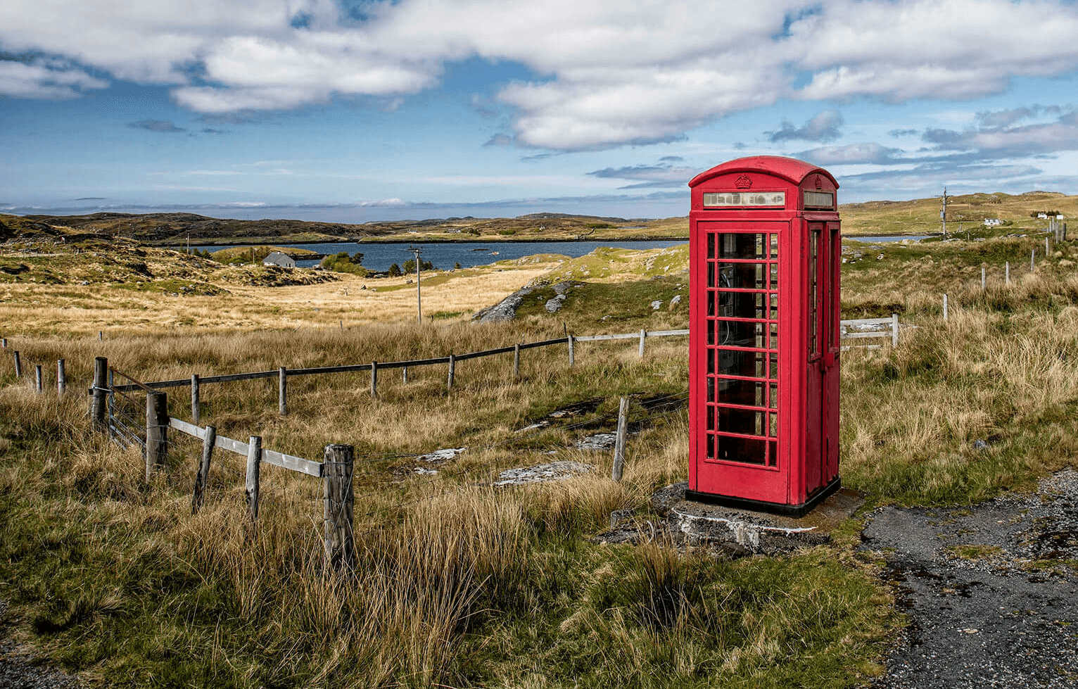 A classic red British phone booth stands alone in a rural landscape of grassy fields, winding fences, and a gravel road under a partly cloudy sky. A small body of water and rolling hills stretch out in the background, highlighting the booth's striking contrast against the natural surroundings.