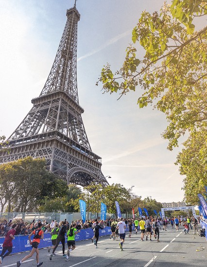 Des coureurs participant à une course sous la Tour Eiffel, avec des barrières de sécurité et des drapeaux bleus tout autour, et une belle journée ensoleillée.