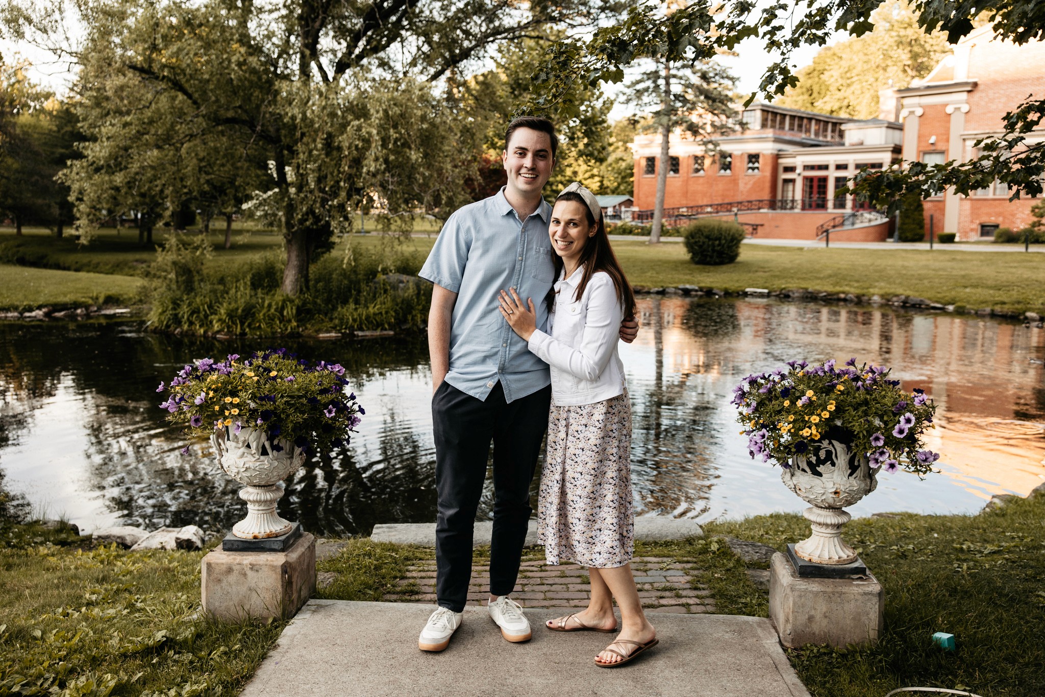 Newly engaged couple standing together on the terrace at Congress Park in Saratoga Springs, she shows off her ring as they smile at the camera — Lizz Spano Photography, Saratoga Springs engagement photographer