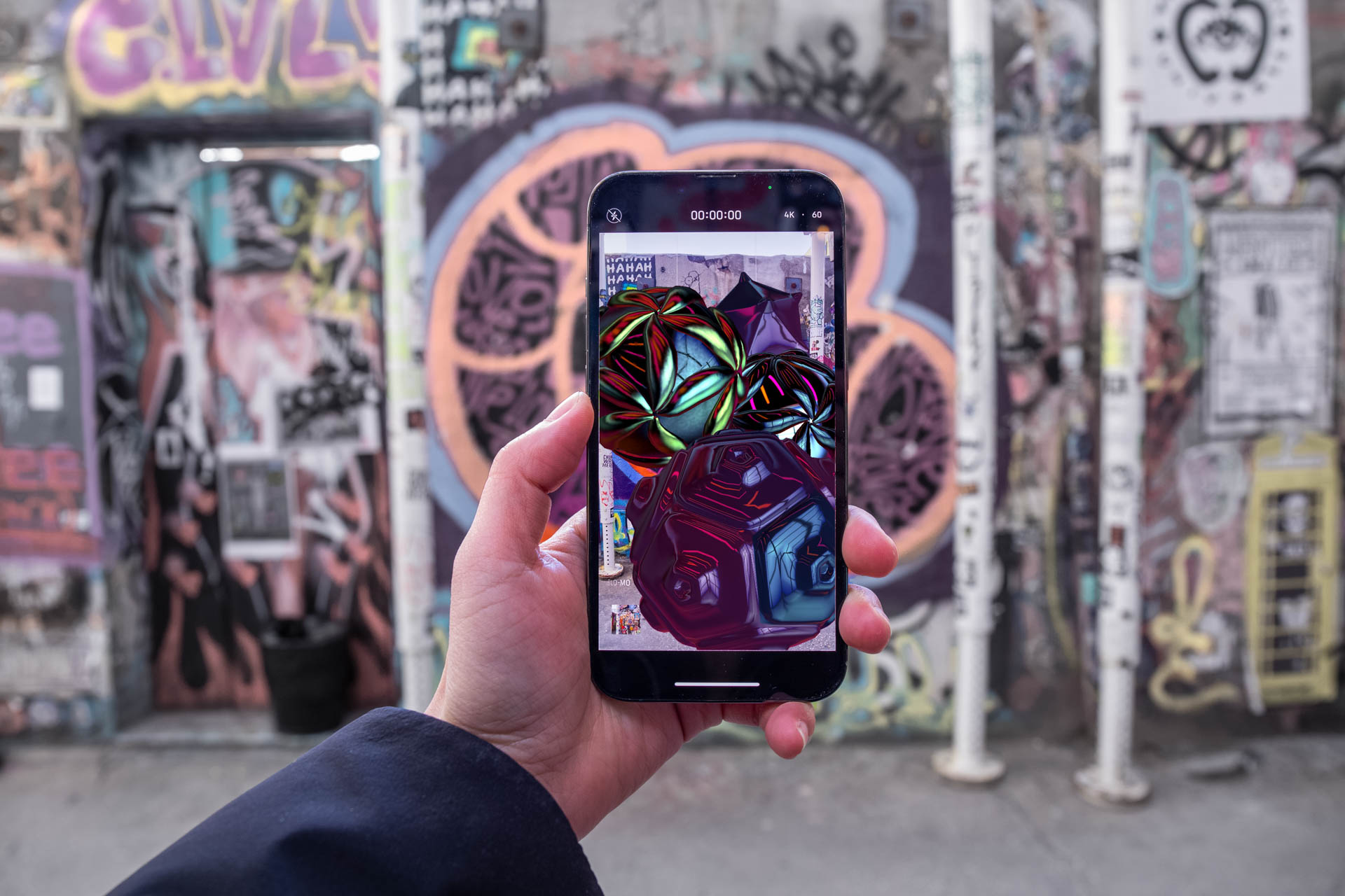 A hand holds a smartphone displaying a colorful, abstract image with swirling patterns in shades of green, red, blue, and purple. The screen shows a digital clock reading "00:00:00." The background features a graffiti-covered wall with vertical poles and various posters.