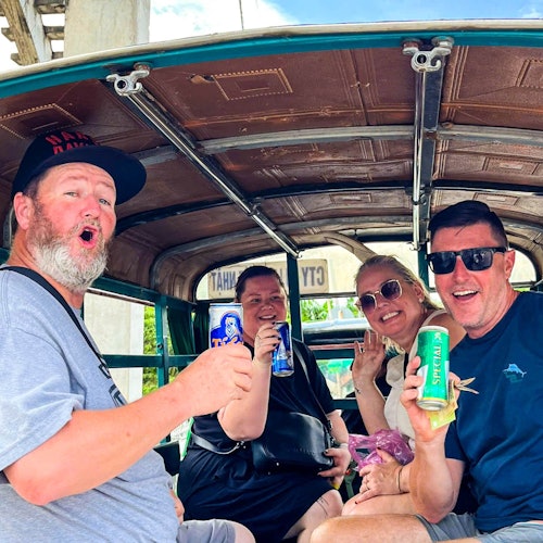 Empat orang duduk di dalam kendaraan terbuka, tersenyum dan memegang minuman. Kendaraan tersebut memiliki atap kayu dan rangka logam.