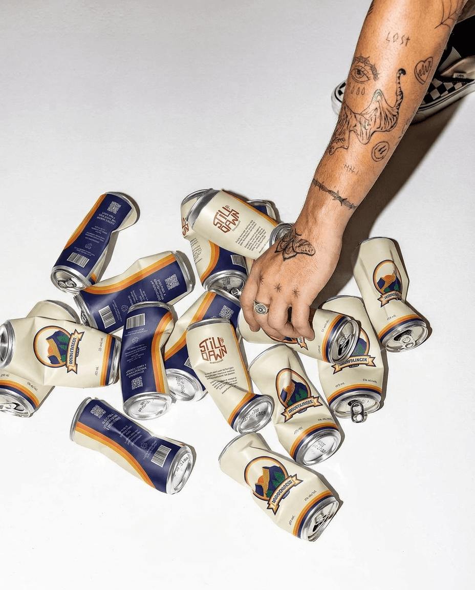 Hand arranging Still Dawn and Mountaineer Lager cans on a white surface.