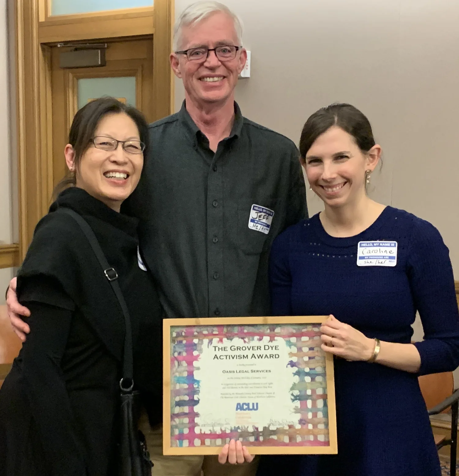Três pessoas sorriem enquanto seguram um prêmio emoldurado intitulado "Prêmio de Ativismo Grover Dye" em uma sala com iluminação acolhedora.