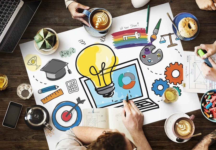 People brainstorming around a table with drawings of a light bulb, laptop, and creative icons on paper.