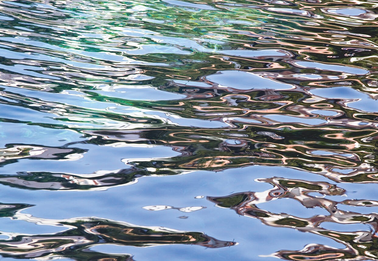 Fein gekräuselte Wasseroberfläche, in der sich blauer Himmel und grüne Ufer spiegeln