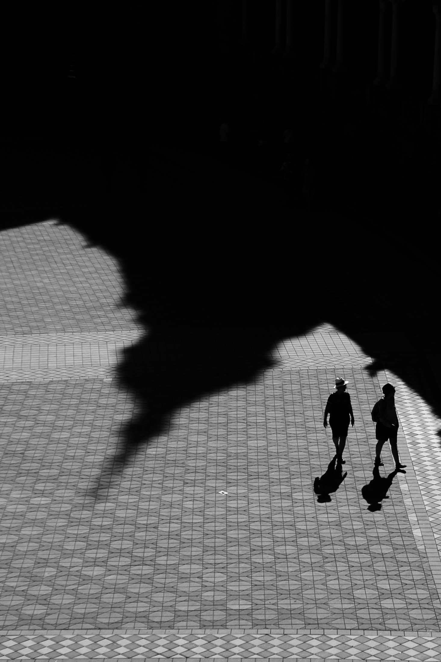 Two people walking across a large square covered by dramatic shadow, minimalist black and white street photography.