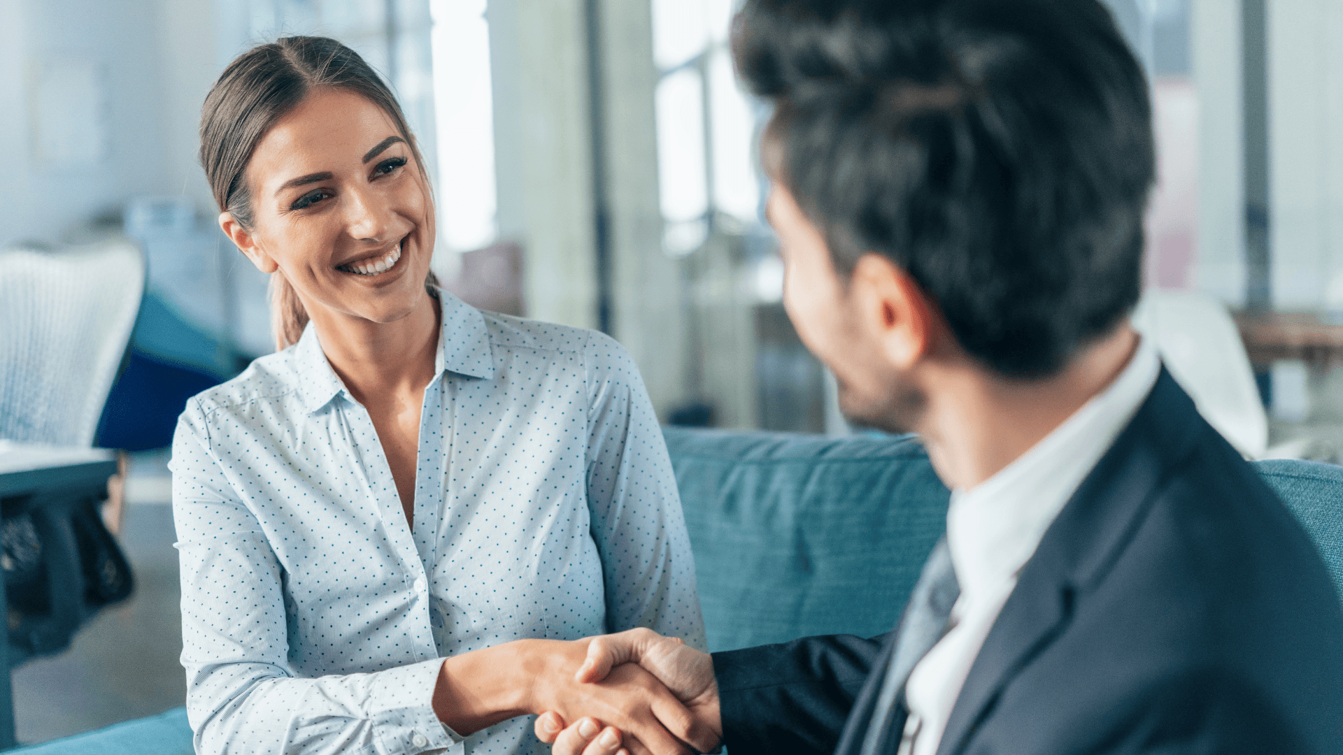 Frau blickt lächelnd einen Mann an und gibt ihm die Hand, vom Mann ist nur der Hinterkopf, schwarze Haare zu sehen, beide sind businessmäßig gekleidet mit Bluse und Hemd und Krawatte
