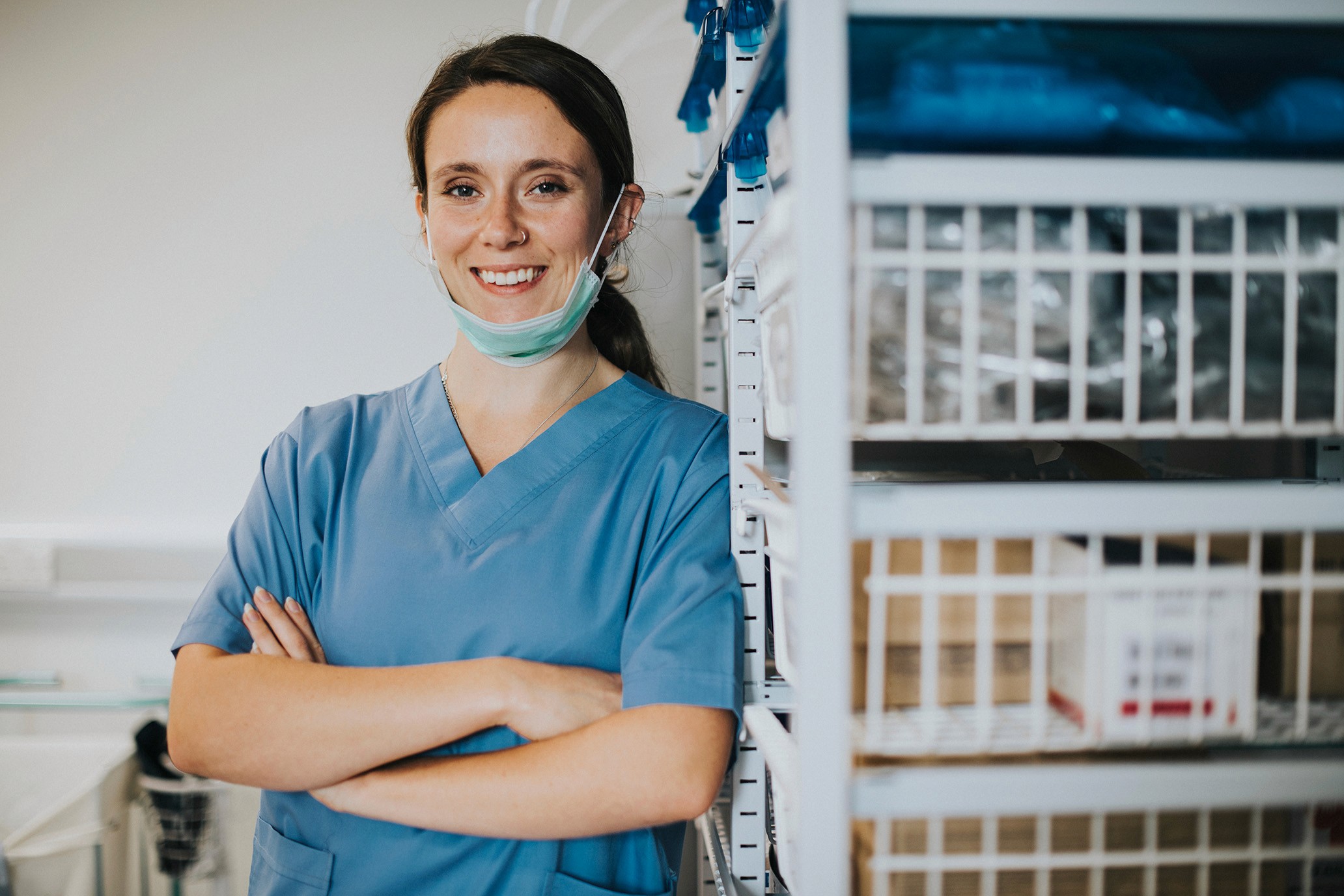 Smiling medical professional with arms crossed, wearing scrubs and a face mask, standing beside storage shelves.