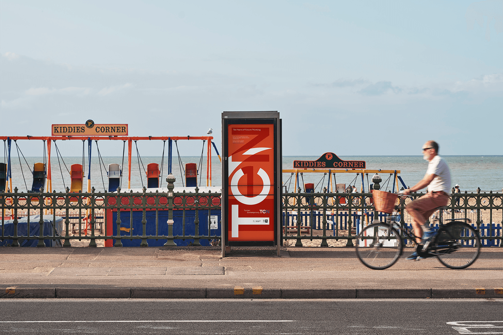 A person rides a bicycle past a brightly colored poster along a seaside promenade.