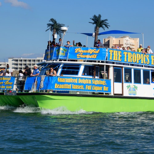 Bright green and blue tour boat named "The Tropics" with passengers on board, cruising on water under a clear sky.