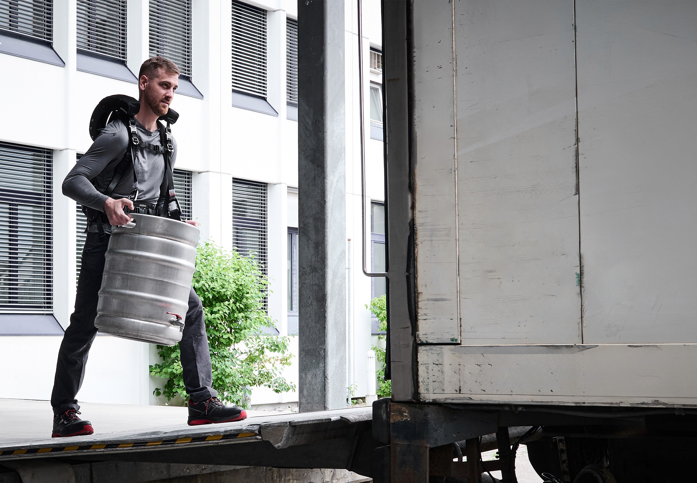 A worker wearing the CarrySuit exoskeleton lifts a metal barrel to load it into a truck.