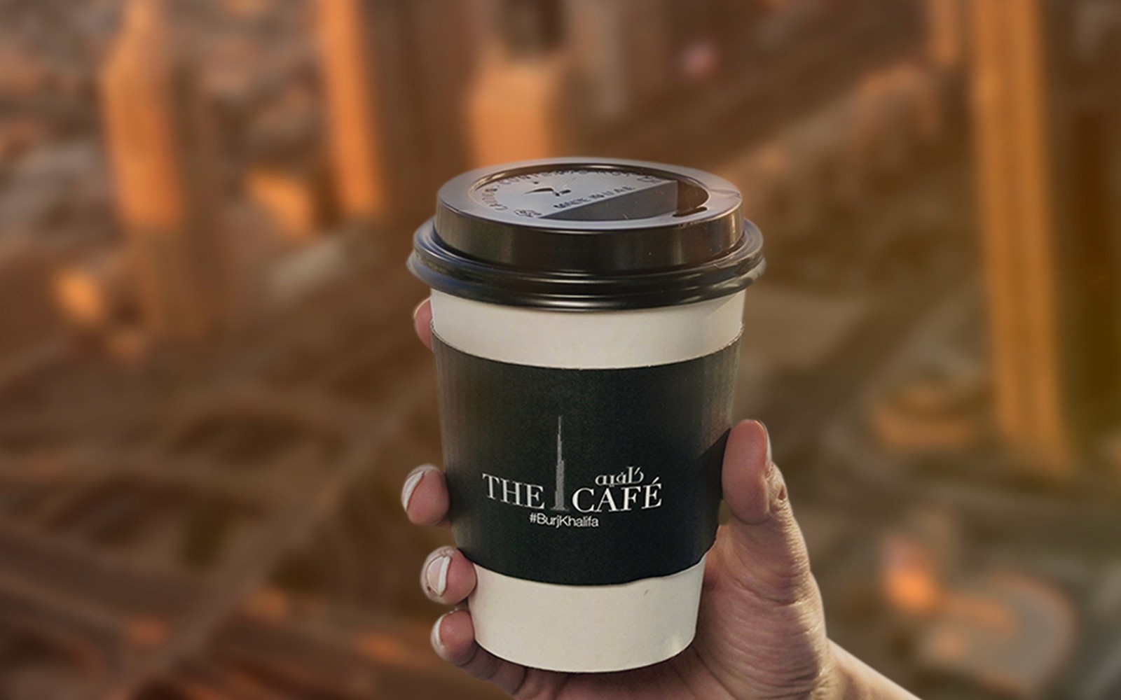 Ruka držící šálek kávy z kavárny The Café v Burj Khalifa s výhledem na město v pozadí.
