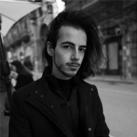A young person with long hair and a beard, wearing a dark coat, stands on a city street with historic architecture in the background on a cloudy day.