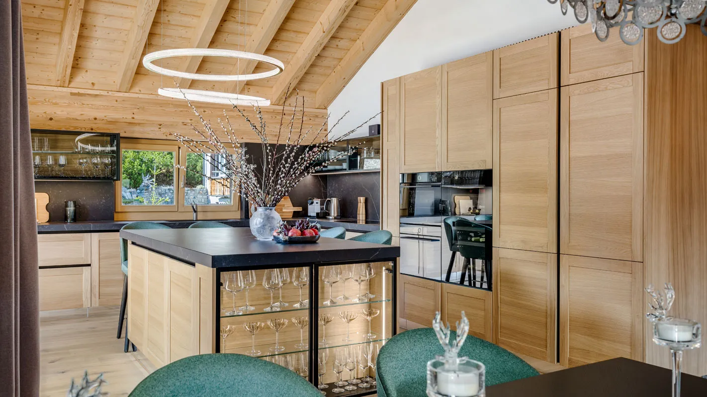 Silvretta Klosters chalet kitchen with central island, glass-front storage, and light oak cabinetry under timber ceiling