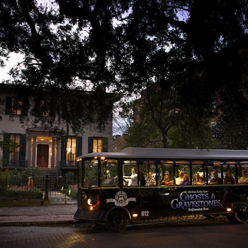 傍晚时分,一辆标有“幽灵与墓碑”字样的旅游巴士停在街上,里面载有乘客,背景是一座灯火通明的大房子。