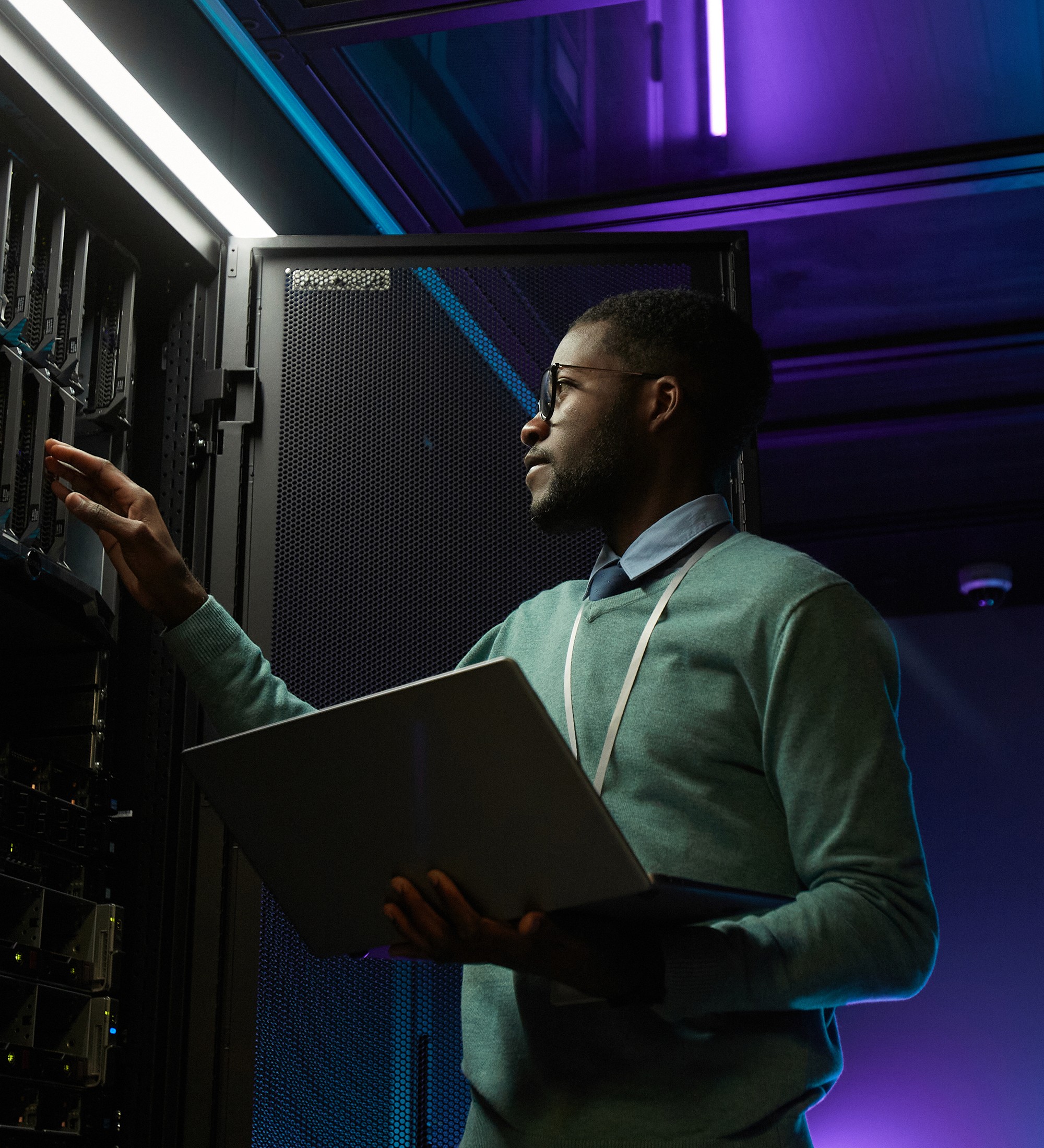 A person in a mint-colored sweater stands in what appears to be a server room or data center, holding a laptop. They are gesturing with one hand while surrounded by purple and blue lighting and technical equipment.