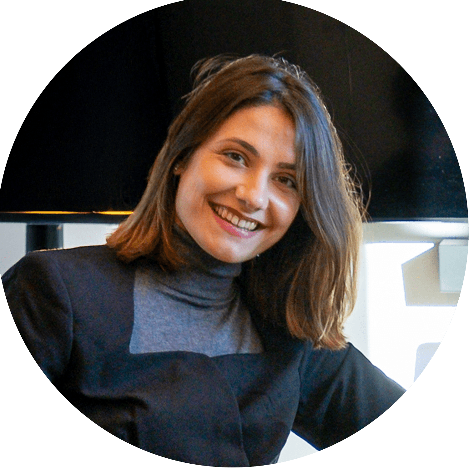Smiling woman with medium-length brown hair, wearing a gray top and black apron, indoors. Background features soft lighting, conveying a warm, welcoming mood.
