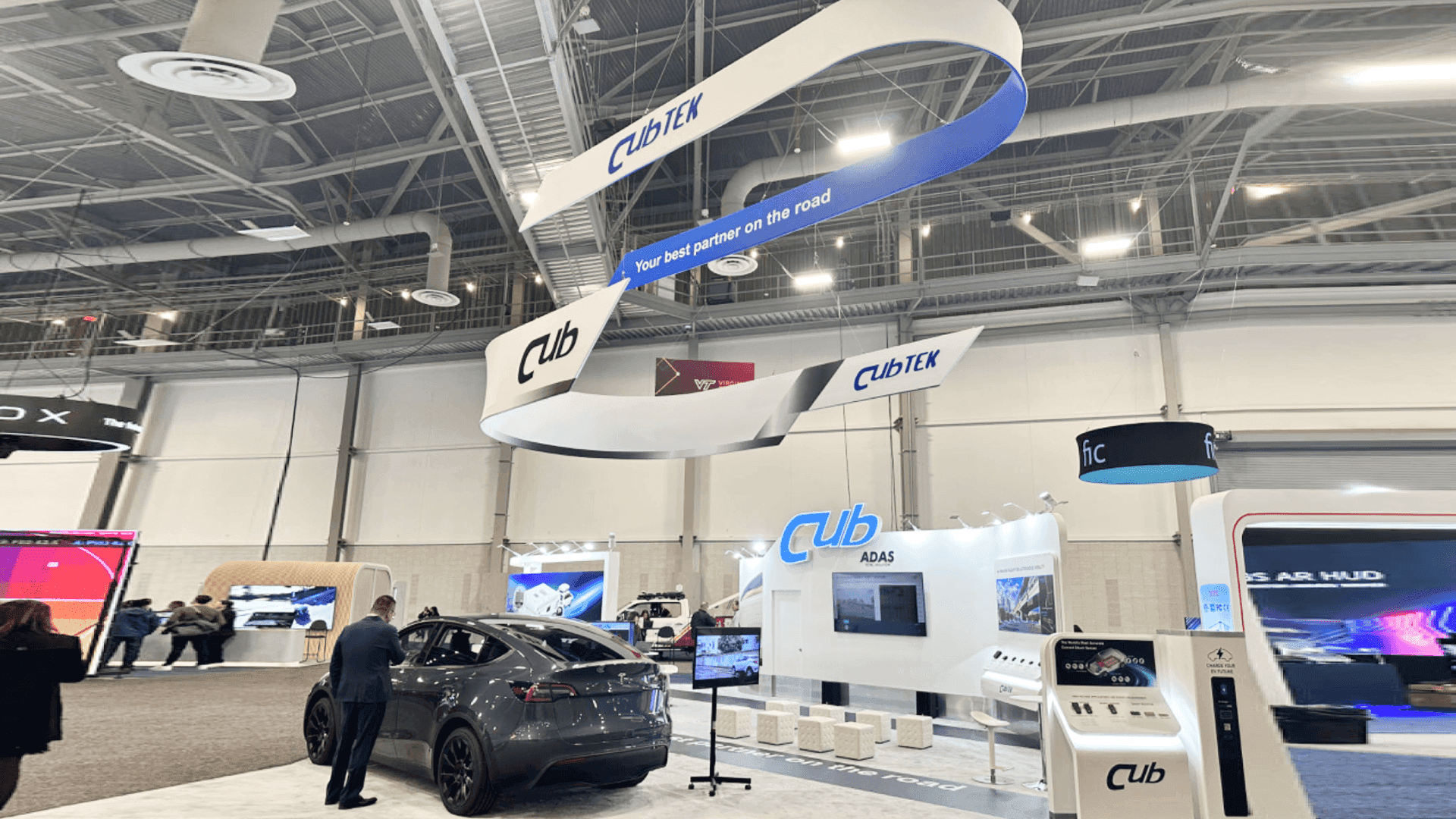 CUB-TEK CES 2024 large island vehicle and equipment display booth with suspended curved banner elements, car demo zone, product monitor, and open visitor circulation space.