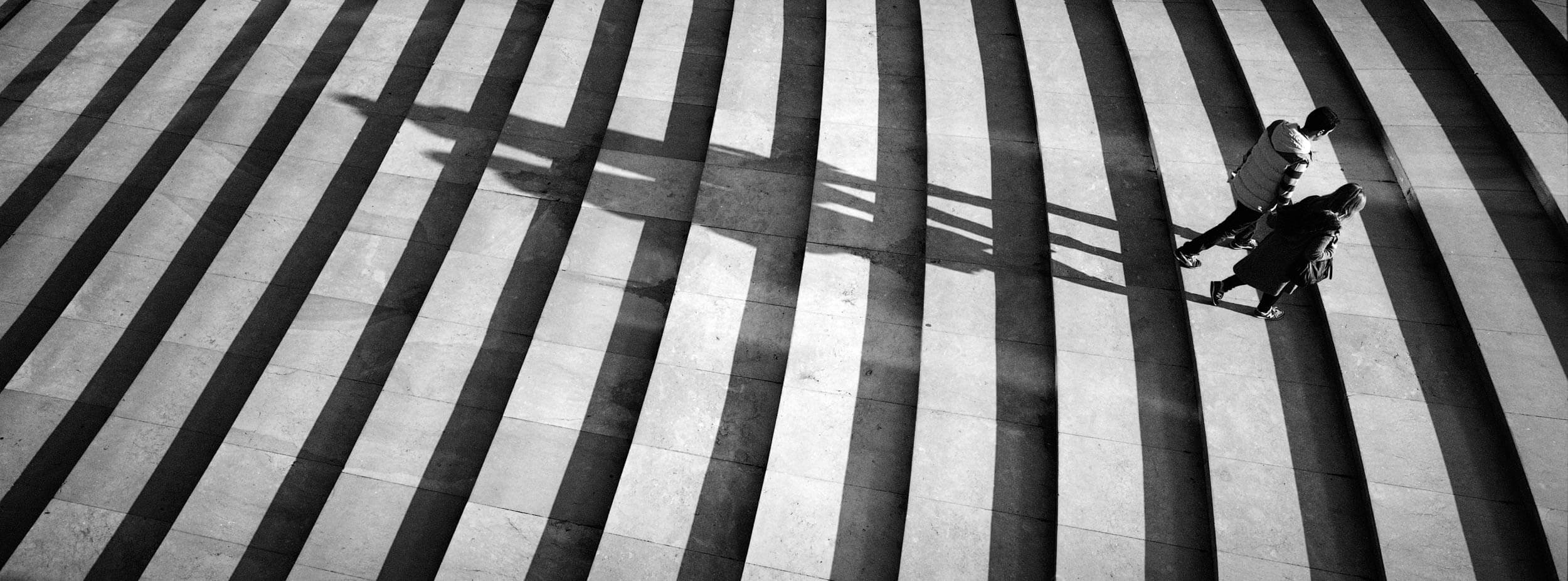 Black and white stair with 2 people walking up casting long shadows behind them shot on the Hasseblad XPan  