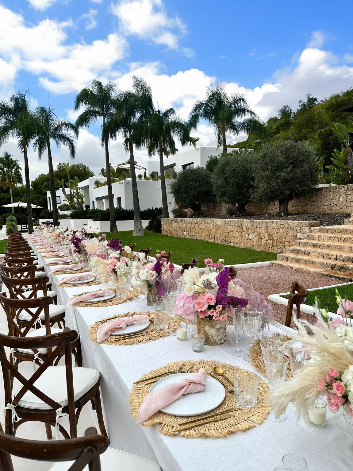 Long outdoor event table with floral arrangements and place settings on the estate lawn near stone steps.