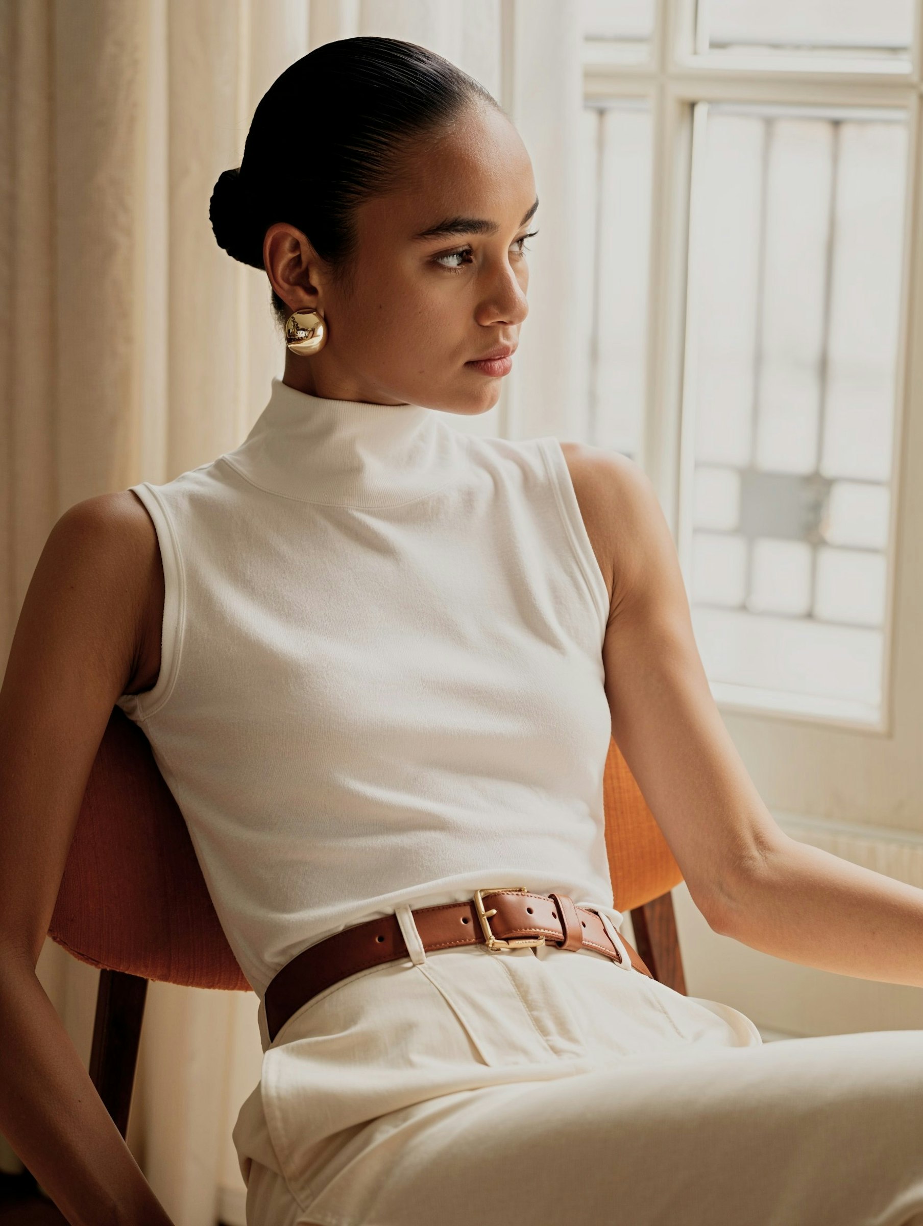 Elegant woman in white turtleneck and cream trousers, accessorized with gold earrings and a brown belt, sitting by a softly lit window.
