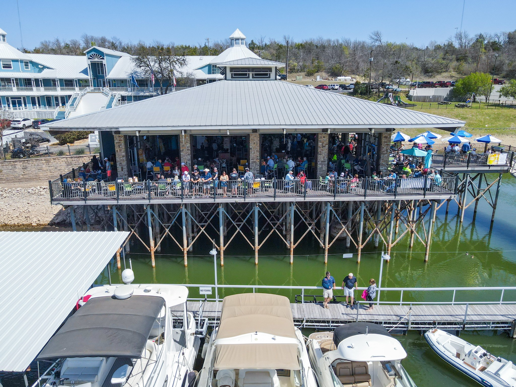 The image shows a bustling waterfront restaurant on stilts with a large gathering of people, overlooking a marina filled with boats under a clear blue sky.