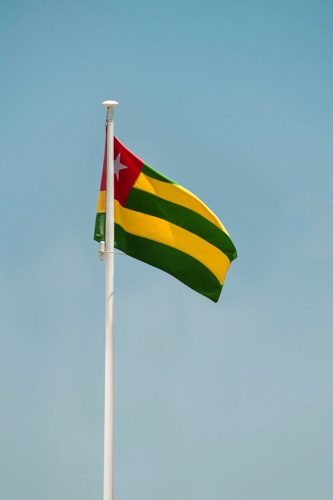 The flag of Togo waving in the clear blue sky, featuring its distinctive design with five horizontal stripes in green and yellow, and a red square with a white star in the top left corner.