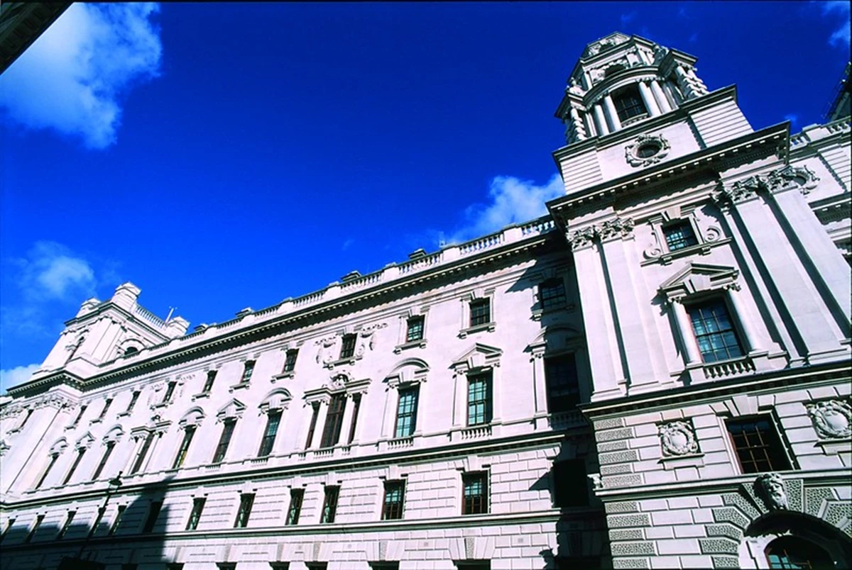A large historic building with ornate stone architecture is shown against a bright blue sky. The perspective looks upward along the façade, highlighting towers and detailed carvings.