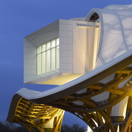 在黄昏的天空下,一个现代建筑结构展示了一个大型发光的白色箱体,从由木材和织物制成的弯曲屋顶延伸出来。