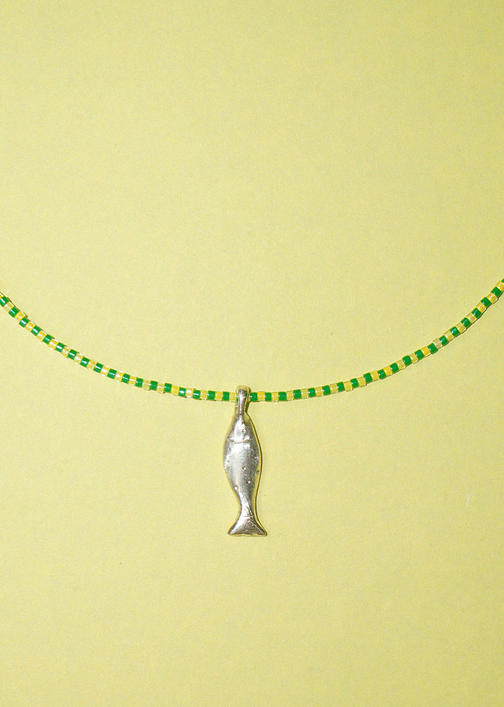 A silver fish-shaped pendant necklace on a green and yellow beaded chain, displayed against a solid pale yellow background.