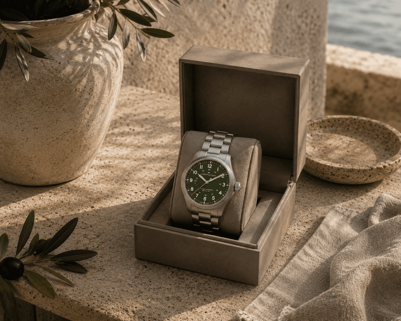 A Hamilton Khaki Field mechanical watch with a stainless steel bracelet and a deep olive green dial displayed inside an open taupe presentation box. The watch sits on a travertine balcony ledge overlooking a tranquil blue sea, styled with a ceramic vase containing olive branches, a small stone dish, a sprig of olive leaves with a black olive, and draped beige linen under soft natural sunlight.